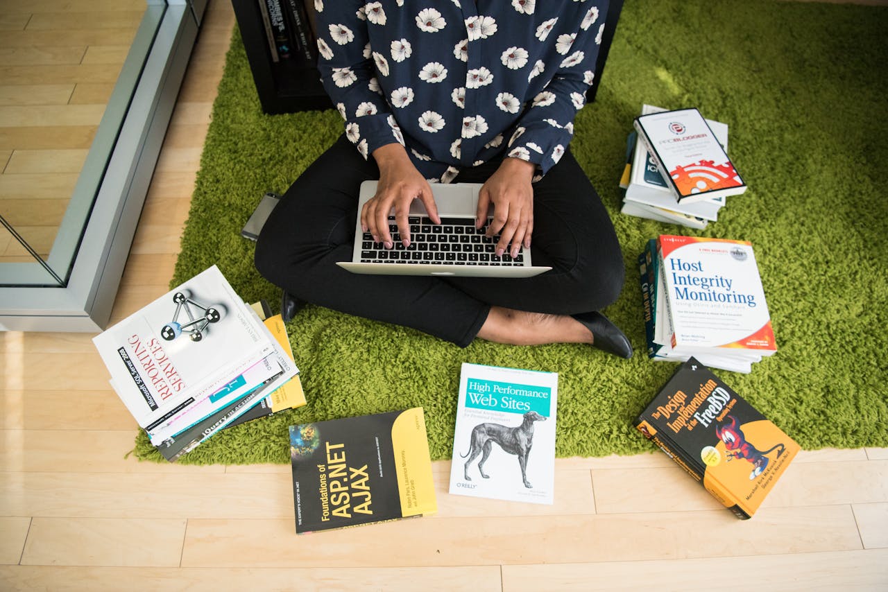 Woman sitting on green rug working on laptop, surrounded by technology books in a modern room.