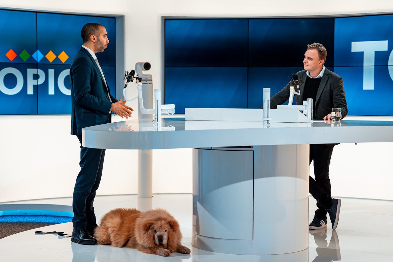 Two hosts discussing at a modern TV studio with a dog laying by. Engaging professional setup.